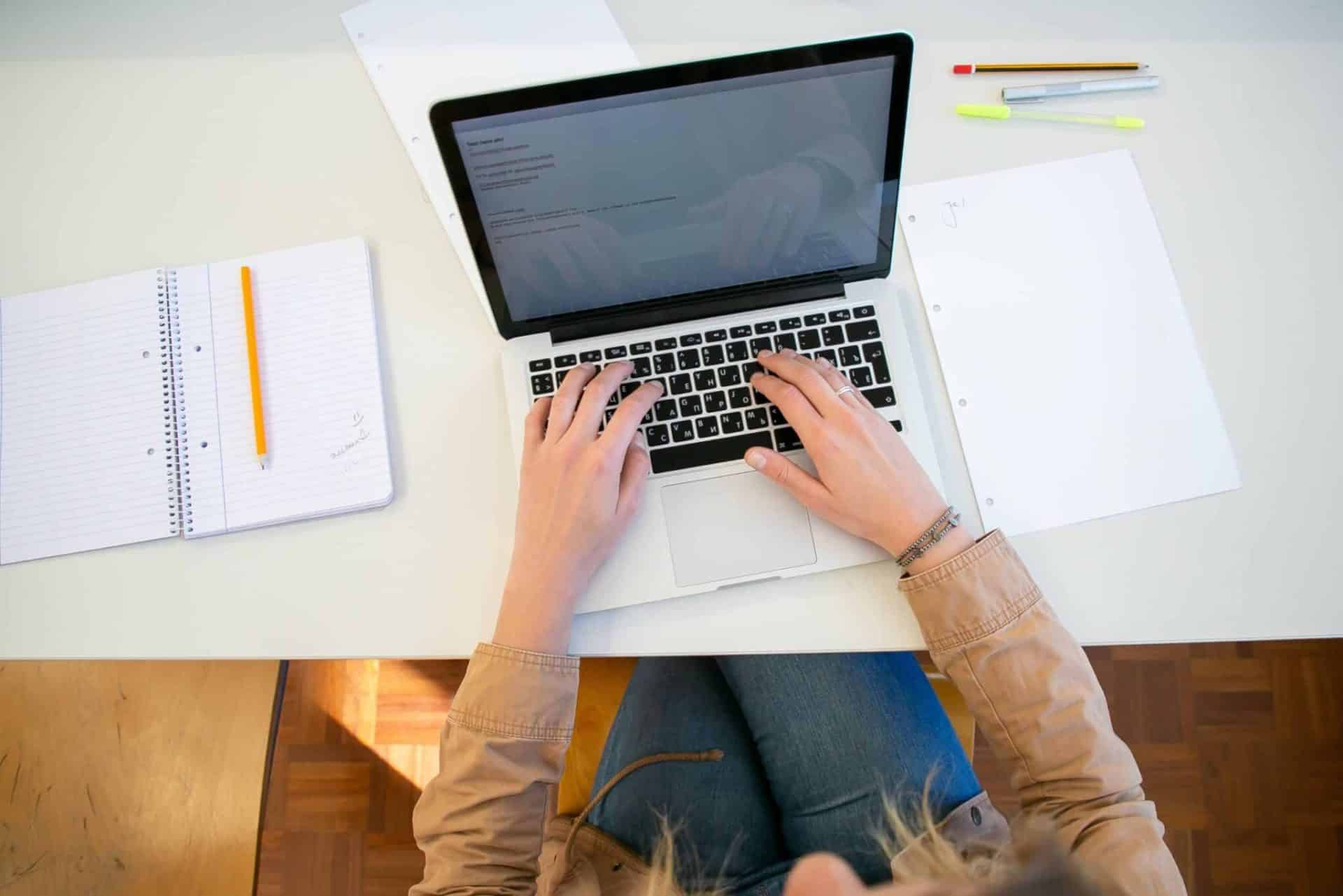 Vue de dessus d'une personne tapant sur un ordinateur portable, entourée de feuilles, d'un cahier et de stylos sur un bureau blanc