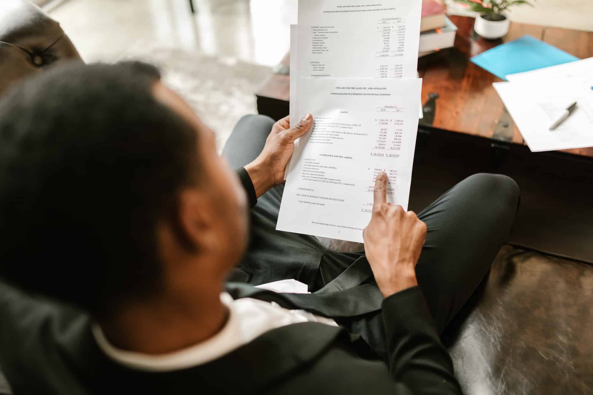 Homme en costume assis, consultant des documents financiers ou comptables, pointant une ligne sur une feuille contenant des chiffres.
