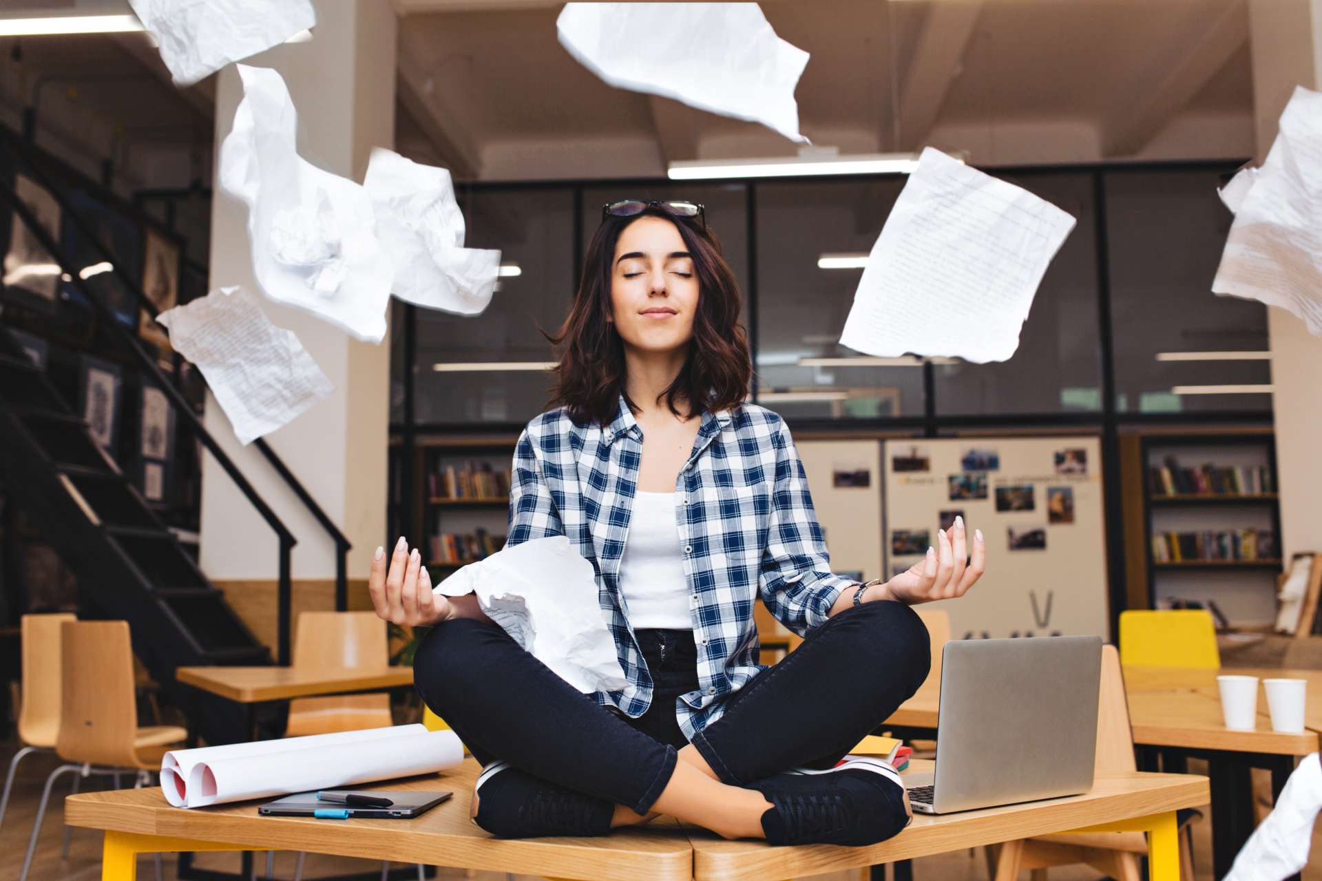 Une femme médite en tailleur sur une table dans un espace de coworking, entourée de feuilles de papier en l'air.